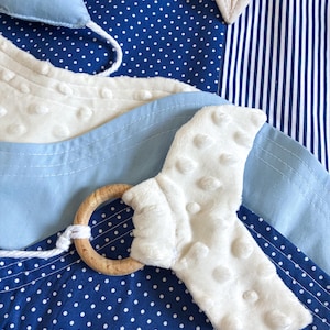May include: A baby toy with a blue and white color scheme. The toy features a wooden ring, a white textured fabric, and a blue fabric with white polka dots. The background includes blue and white striped fabric.
