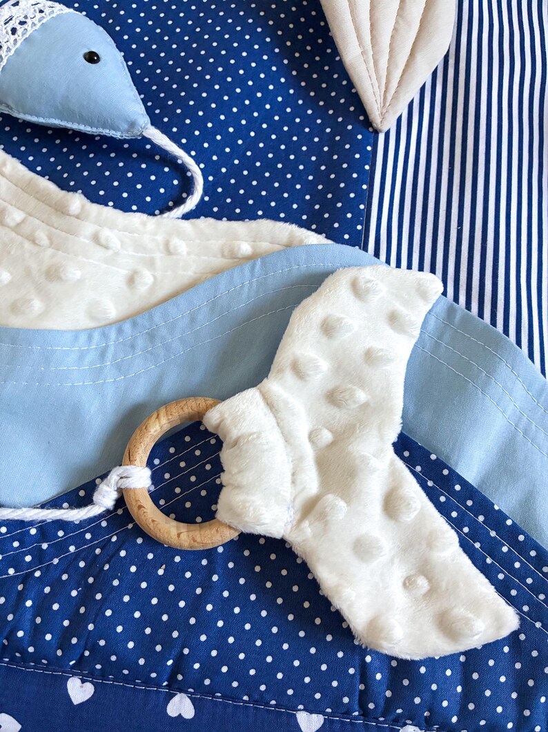 May include: A baby toy with a blue and white color scheme. The toy features a wooden ring, a white textured fabric, and a blue fabric with white polka dots. The background includes blue and white striped fabric.