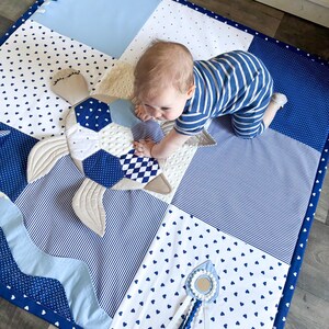 May include: A patchwork baby play mat in shades of blue and white, featuring a turtle-shaped toy. The mat has various patterns, including hearts, stripes, and polka dots. A small mirror toy with ribbons is also visible.