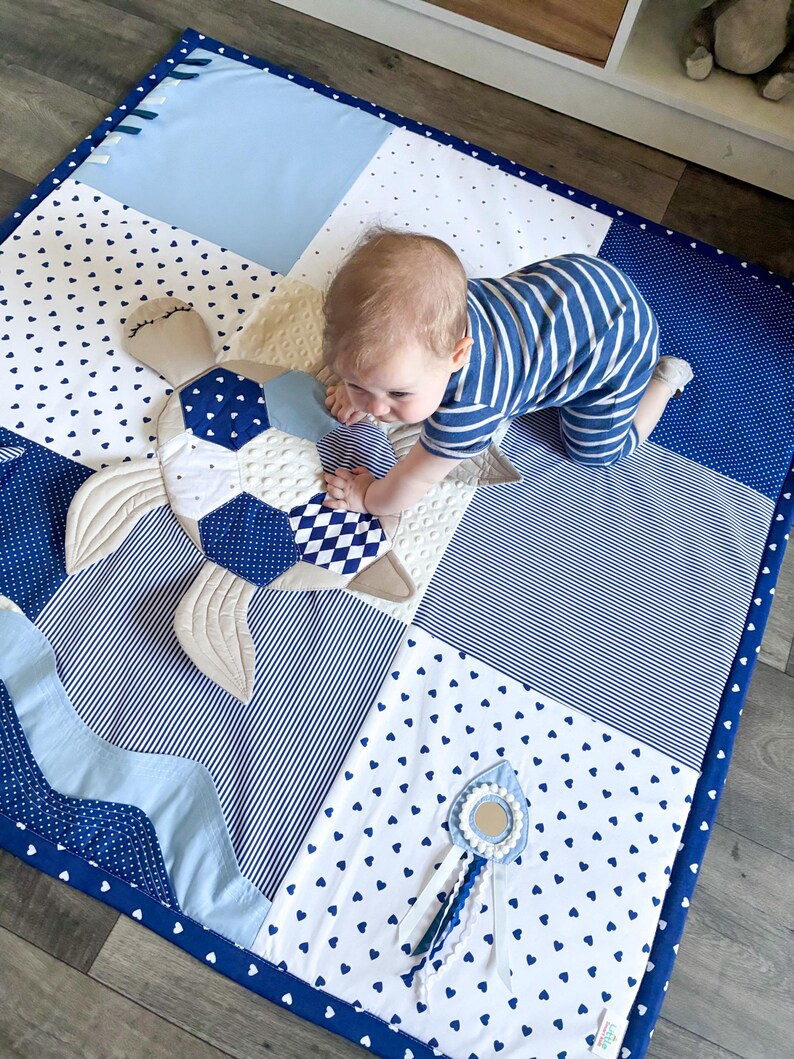 May include: A patchwork baby play mat in shades of blue and white, featuring a turtle-shaped toy. The mat has various patterns, including hearts, stripes, and polka dots. A small mirror toy with ribbons is also visible.