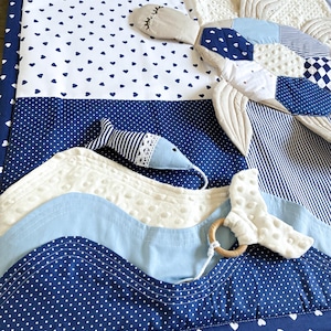May include: A baby play mat with a nautical theme. The mat features various shades of blue and white fabrics, including polka dots, hearts, and stripes. A stuffed whale and fish are attached, along with a wooden teething ring.