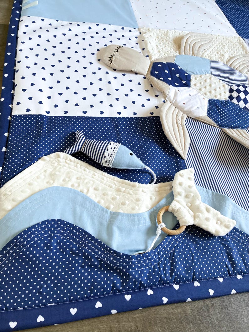 May include: A baby play mat with a nautical theme. The mat features various shades of blue and white fabrics, including polka dots, hearts, and stripes. A stuffed whale and fish are attached, along with a wooden teething ring.