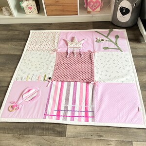 May include: A pink and white patchwork play mat with various textures and patterns. Features include a crown, a rattle, and a floral branch. The mat is designed for babies and toddlers.