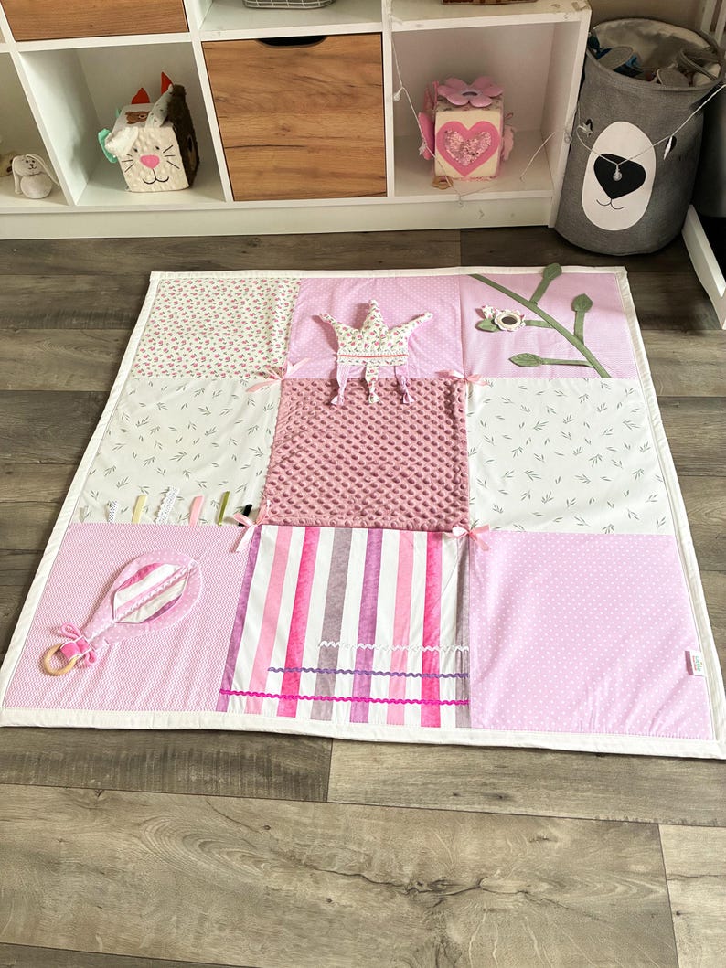 May include: A pink and white patchwork play mat with various textures and patterns. Features include a crown, a rattle, and a floral branch. The mat is designed for babies and toddlers.