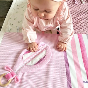 May include: A pink baby play mat with a variety of textures and patterns, including a chevron design and striped sections. A soft, pink toy with a wooden ring is attached. The mat is designed for infant play and development.