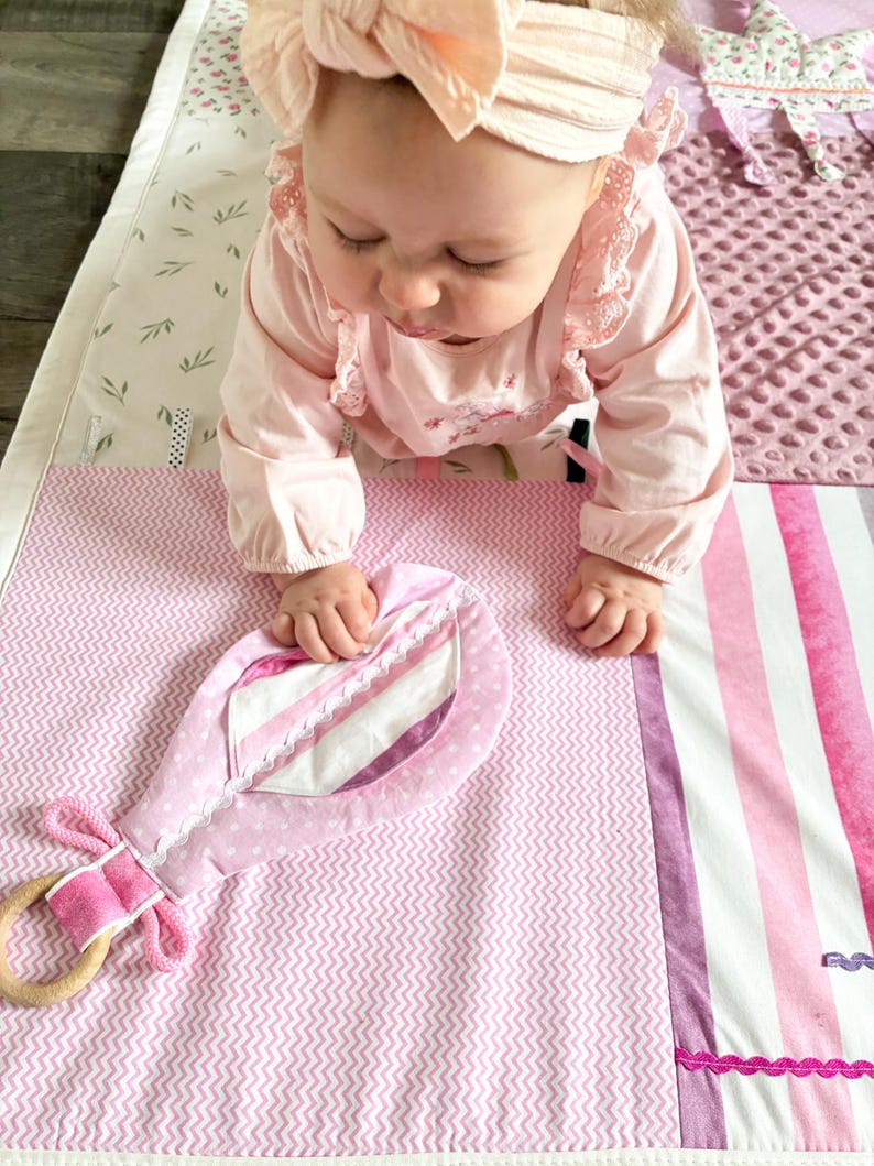 May include: A pink baby play mat with a variety of textures and patterns, including a chevron design and striped sections. A soft, pink toy with a wooden ring is attached. The mat is designed for infant play and development.