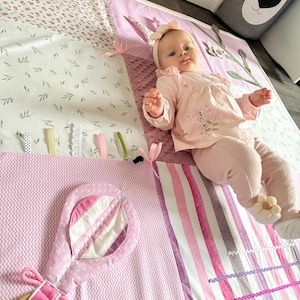 May include: A baby girl wearing a pink outfit and headband lies on a soft, colorful play mat. The mat features floral, leaf, and striped patterns in shades of pink and white, with a pink rattle toy. The mat has ribbons and textures for sensory play.