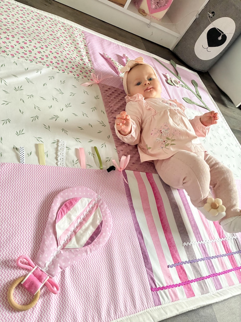 May include: A baby girl wearing a pink outfit and headband lies on a soft, colorful play mat. The mat features floral, leaf, and striped patterns in shades of pink and white, with a pink rattle toy. The mat has ribbons and textures for sensory play.