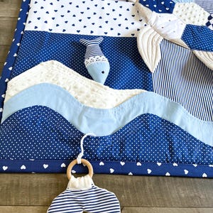 May include: A nautical-themed baby play mat with a whale tail teether. The mat features blue and white fabrics with heart and polka dot patterns, and wave-like appliqu&eacute;s. A stuffed fish and a whale-shaped pillow are also present, creating a sea-inspired nursery item.