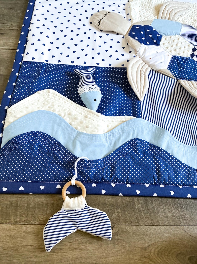 May include: A nautical-themed baby play mat with a whale tail teether. The mat features blue and white fabrics with heart and polka dot patterns, and wave-like appliqu&eacute;s. A stuffed fish and a whale-shaped pillow are also present, creating a sea-inspired nursery item.