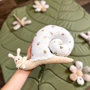 May include: A plush snail toy with a white, floral-patterned shell and a cream-colored body. The snail has a cream-colored bow and is held over a green leaf-shaped play mat with flower and dragonfly accents. A whimsical children's toy.