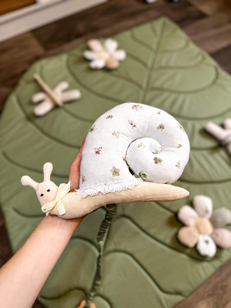 May include: A plush snail toy with a white, floral-patterned shell and a cream-colored body. The snail has a cream-colored bow and is held over a green leaf-shaped play mat with flower and dragonfly accents. A whimsical children's toy.