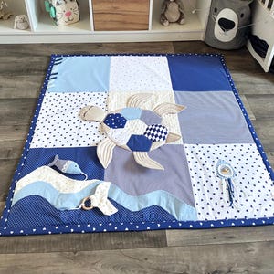 May include: A patchwork baby play mat in shades of blue and white. The mat features a turtle applique, a fish, and a wave design. The border is a solid dark blue with white polka dots. The mat measures approximately 36 inches square.