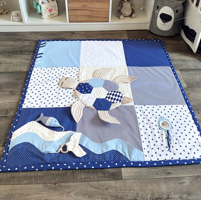 May include: A patchwork baby play mat in shades of blue and white. The mat features a turtle applique, a fish, and a wave design. The border is a solid dark blue with white polka dots. The mat measures approximately 36 inches square.