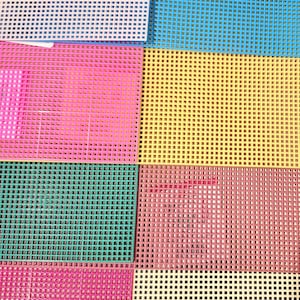 May include: A collection of rectangular plastic canvas sheets in various colors including white, blue, yellow, pink, teal, and light yellow. Each sheet features a uniform grid of square holes for crafting projects, with a product label on the white sheet.