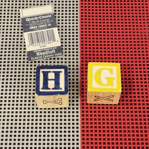 May include: Two wooden alphabet blocks featuring the letters H and G, positioned on a gray and red plastic grid canvas. The block with the letter H has a blue border, while the block with the letter G has a yellow border. A Quick-Count label is included.