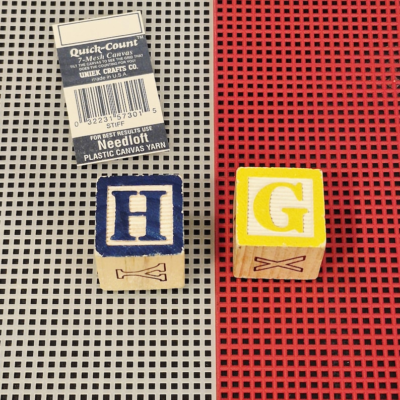 May include: Two wooden alphabet blocks featuring the letters H and G, positioned on a gray and red plastic grid canvas. The block with the letter H has a blue border, while the block with the letter G has a yellow border. A Quick-Count label is included.