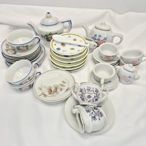 May include: A collection of miniature porcelain tea party set pieces. The set includes teacups, saucers, teapots, a sugar bowl, and plates. The pieces are white with floral and pine cone designs, and some have blue trim. The plates measure approximately 4 inches in diameter.