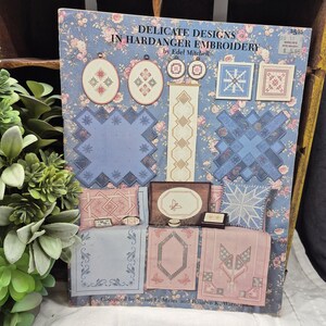May include: A book titled "Delicate Designs in Hardanger Embroidery" by Edel Mitchell. The cover features various cross-stitch patterns in pink, blue, and white, including geometric shapes and floral designs. The background is a blue floral pattern.