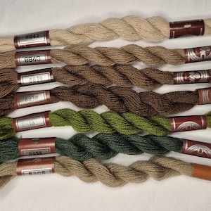 May include: A collection of embroidery floss in various shades of brown and green. Each skein is labeled with a number and wrapped with a brown paper band. The floss is arranged in a neat, parallel row, showcasing the different colors and textures.