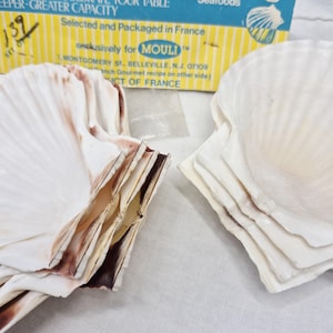 May include: Close-up of several white scallop shells with brown and tan accents. The shells are stacked and arranged near a box with text that reads "Selected and Packaged in France."