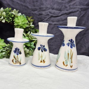 May include: Three ceramic candle holders with a white base, blue trim, and hand-painted blue iris flower designs. The candle holders are different sizes and shapes, with a classic, elegant style.