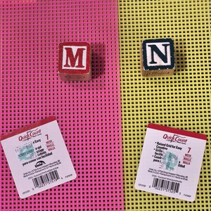 May include: Two square plastic mesh sheets in fluorescent pink and yellow, each with a white product label. Two wooden alphabet blocks, one red with the letter M and one black with the letter N, are placed on the respective mesh surfaces.