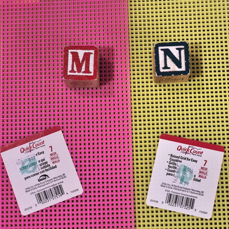May include: Two square plastic mesh sheets in fluorescent pink and yellow, each with a white product label. Two wooden alphabet blocks, one red with the letter M and one black with the letter N, are placed on the respective mesh surfaces.