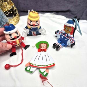 May include: Four wooden hanging ornaments featuring two egg-shaped figures with mustaches, a doll with red yarn hair in a blue and white plaid outfit, and a flat figure wearing a green hat and white dress with red and green holly accents.