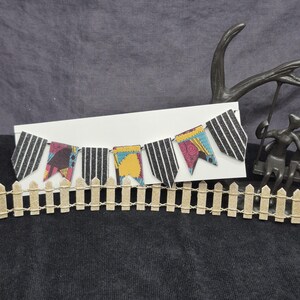 May include: A decorative banner of small, colorful pennants with black and white striped and patterned designs. The banner is displayed on a white card, with a miniature wooden fence in the foreground. The background is a dark gray fabric.