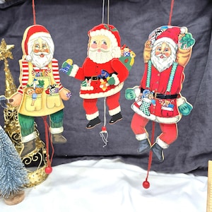 May include: Three hanging Santa Claus ornaments made of wood, each featuring a different design with red, white, and green colors. These articulated figures have strings with red beads at the bottom, allowing the arms and legs to move when pulled.
