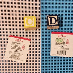 May include: Two sheets of plastic canvas, one pastel pink and one bright blue, each featuring a 7-mesh grid. Two wooden alphabet blocks are present: one yellow block with the letter C and one dark blue block with the letter D. Two product labels.