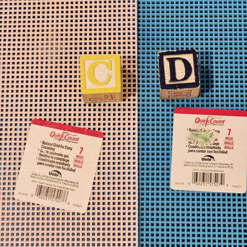 May include: Two sheets of plastic canvas, one pastel pink and one bright blue, each featuring a 7-mesh grid. Two wooden alphabet blocks are present: one yellow block with the letter C and one dark blue block with the letter D. Two product labels.
