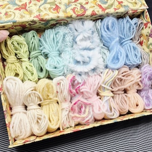 May include: A vintage box filled with colorful yarn skeins in shades of green, blue, pink, and cream. The box has a decorative floral pattern on the lid and interior. The yarn is bundled and tied, perfect for crafting projects.