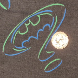 May include: Black fabric with a Batman logo in blue and green outline. A U.S. quarter coin is next to the logo for size comparison. The Batman symbol is a stylized bat silhouette.
