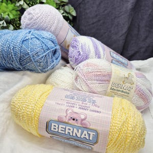 May include: A collection of Bernat Baby Coordinates yarn skeins in various colors, including yellow, blue, white, and pink. The yarn is labeled as sports weight, with a weight of 6 oz. / 170g. The packaging features a teddy bear graphic.