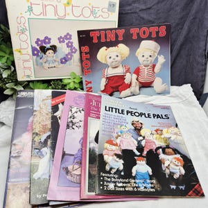 May include: A collection of vintage sewing patterns and books, including "Tiny Tots" and "Little People Pals." The books feature illustrations of dolls and instructions for creating them. The color palette includes pink, white, and red.