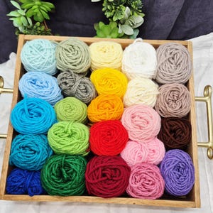 May include: A wooden tray filled with colorful yarn balls in various shades of blue, green, yellow, orange, pink, red, brown, and purple. The yarn is neatly arranged, suggesting a craft or knitting project. The tray has gold-colored handles.