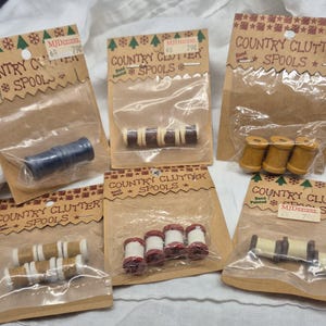 May include: Several packages of hand-painted wooden spools. The spools are in various colors, including blue, brown, red, and yellow, and are packaged on brown cardboard with the text "COUNTRY CLUTTER SPOOLS".