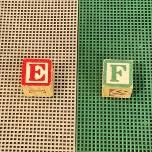 May include: Two wooden alphabet blocks. One block features a red letter E on a white background with a black letter I on the side. The other block features a green letter F on a white background with a black letter W on the side. Both are square.