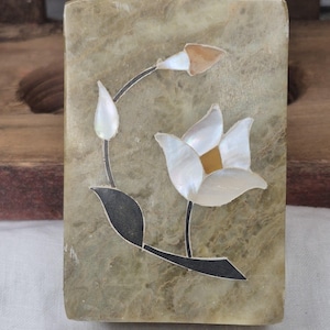 May include: A rectangular, marble-like box with a floral inlay. The flower features white petals with a yellow center, a brown bud, and black and gray leaves. The box has a mottled, light green and brown color scheme.