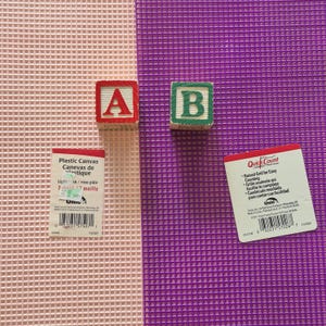 May include: Two sheets of plastic canvas, one light pink and one bright purple, each featuring a grid pattern. Two wooden alphabet blocks, one with a red letter A and one with a green letter B, are placed on the sheets alongside two product labels.