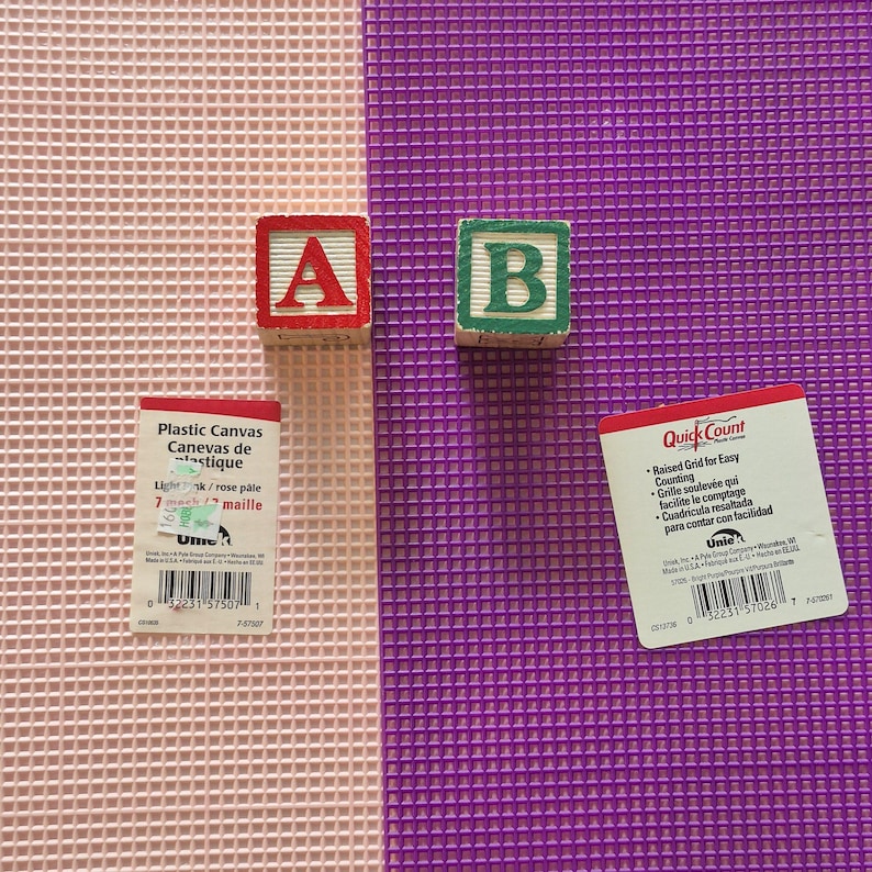 May include: Two sheets of plastic canvas, one light pink and one bright purple, each featuring a grid pattern. Two wooden alphabet blocks, one with a red letter A and one with a green letter B, are placed on the sheets alongside two product labels.