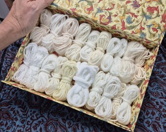 May include: A box filled with bundles of white and off-white yarn. The box has a floral pattern on the lid and is being held open. The yarn is neatly arranged, suggesting a craft or knitting project.