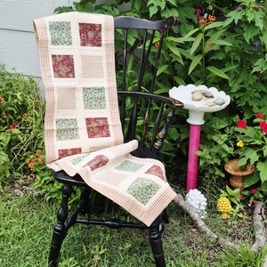 May include: A rectangular table runner featuring a patchwork design of alternating cream squares and squares with red floral or green leaf patterns, bordered by a small red and white gingham check fabric, draped over a black wooden chair.
