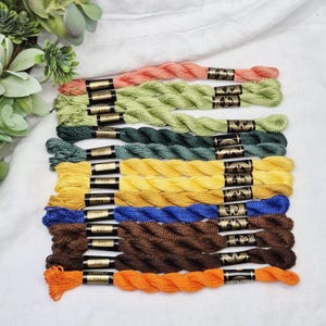 May include: A collection of DMC embroidery floss in various colors, including coral, green, yellow, blue, brown, and orange. Each skein is labeled with a black and gold band. The floss is arranged on a white surface with greenery in the background.