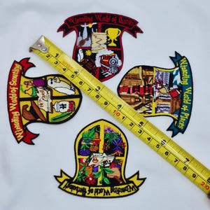 May include: Four embroidered patches with the text "Wizarding World" and various sub-themes. The patches are in the shape of crests, with detailed images and text. A yellow tape measure is laid across the patches, showing measurements in inches.