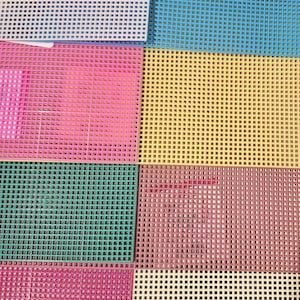 May include: A collection of rectangular plastic canvas sheets featuring a grid of square holes. The sheets are available in various solid colors including white, blue, pink, yellow, teal, and cream, with a product label attached to the white sheet.