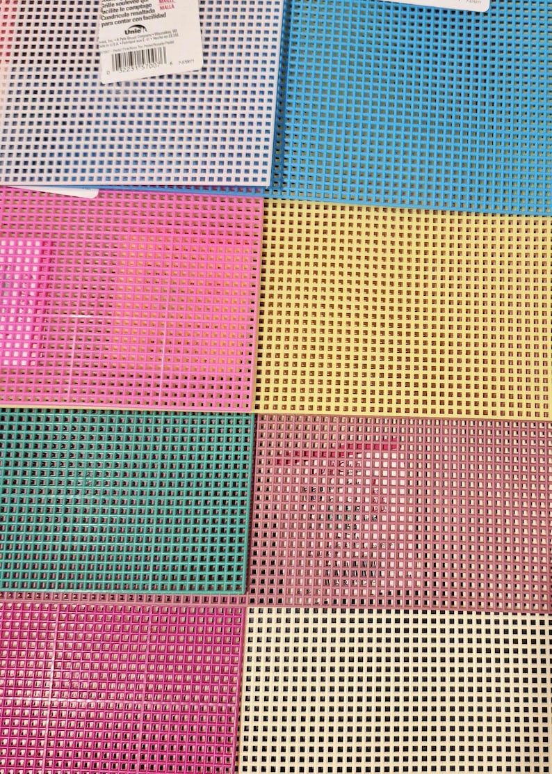 May include: A collection of rectangular plastic canvas sheets featuring a grid of square holes. The sheets are available in various solid colors including white, blue, pink, yellow, teal, and cream, with a product label attached to the white sheet.