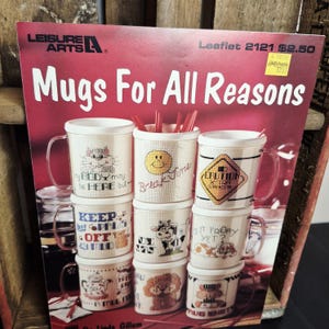 May include: A stack of cream-colored mugs with various cross-stitch designs and text, including a cat, a smiley face, and a caution sign. The mugs are arranged on a red background with the words "Mugs For All Reasons".